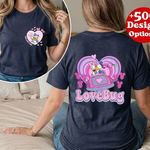 May include: Navy blue t-shirt featuring a heart-shaped graphic with Daisy Duck and Donald Duck. The front of the shirt says "Be My Valentine". The back of the shirt has a "LoveBug" design with Daisy and Donald Duck in a car.