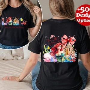 Puede incluir: Camiseta negra con una colorida ilustraci&oacute;n de Ariel de La Sirenita, con un castillo y criaturas marinas. La parte delantera de la camiseta tiene un dise&ntilde;o similar. La camiseta tambi&eacute;n tiene el texto "+500 Design Options".