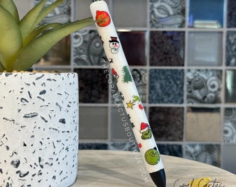 May include: White pen with a black cap and a Christmas themed design. The pen has a variety of Christmas themed stickers including a snowman, a Christmas tree, a Grinch, and a Santa hat. The text "THECORALCACTUSBOUIQUE.COM" is printed on the pen.