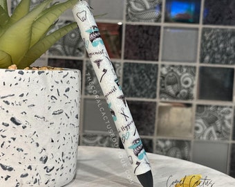 May include: White pen with a blue and black design featuring dental hygiene related icons and the text "Hygienist". The pen is sitting on a white and gray speckled surface.