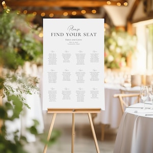 May include: A white wedding seating chart on an easel. The chart reads "Please Find Your Seat" and lists names under table numbers. The wedding date and location are also displayed. The easel is made of wood.