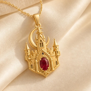 May include: A gold-toned necklace featuring a pendant shaped like a castle with a crescent moon and a star at the top. The center of the castle structure is set with a single, oval-shaped, deep red gemstone. The pendant hangs from a delicate gold chain.