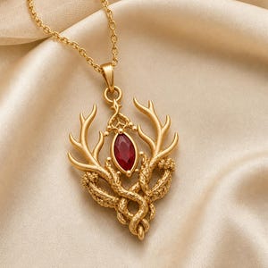 May include: A gold-toned necklace featuring a pendant shaped like a pair of deer antlers intertwined with textured, vine-like elements. A single, marquise-cut deep red gemstone is set vertically in the center of the pendant, suspended from a chain.