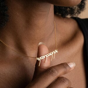 May include: A delicate gold-colored necklace with a script pendant. The pendant features an elegant, stylized inscription. The necklace is worn around the neck, with the pendant resting near the collarbone. The chain is thin and subtle.