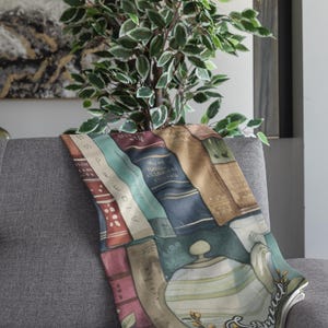 May include: A decorative throw blanket draped over a gray sofa. The blanket features a watercolor design of stacked books, a teapot, and the word "Samuel". A green plant is in the background.