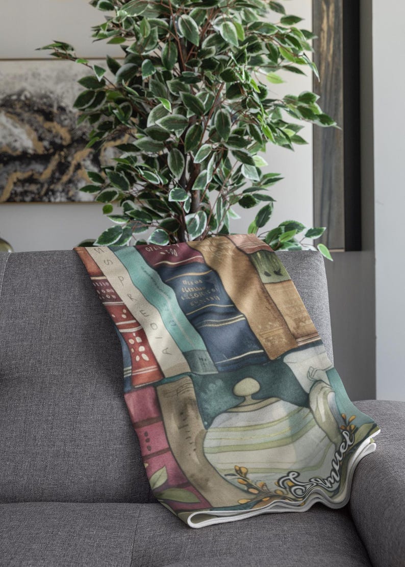 May include: A decorative throw blanket draped over a gray sofa. The blanket features a watercolor design of stacked books, a teapot, and the word "Samuel". A green plant is in the background.
