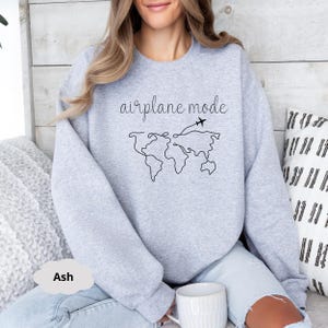 Puede incluir: Una sudadera gris con el texto "airplane mode" y un dibujo lineal del mundo con dos aviones volando sobre &eacute;l. La sudadera la lleva puesta una persona sentada en un sof&aacute;.