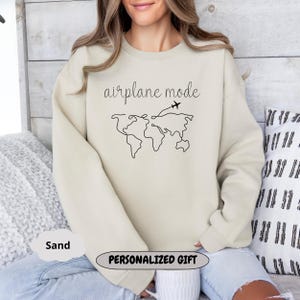 Puede incluir: Una sudadera beige con el texto "modo avi&oacute;n" y un dibujo simple de un mapa del mundo con un avi&oacute;n volando sobre &eacute;l. La sudadera es un regalo personalizado.