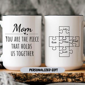 May include: White ceramic coffee mug with a black rim and handle. The mug has a black inscription that reads "Mom You Are The Piece That Holds Us Together". The other side of the mug has a black and white puzzle with the names "Leo", "Susa", "Mom", "Mali", and "Luis" written inside the puzzle pieces. The mug has a white label that reads "Personalized Gift".