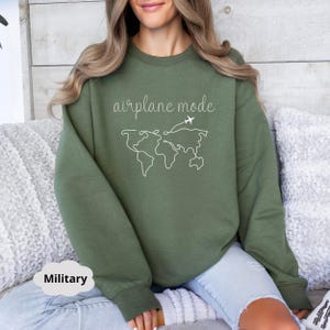 Puede incluir: Una sudadera verde con el texto "modo avi&oacute;n" y un dibujo lineal de un mapa del mundo con aviones volando sobre &eacute;l.