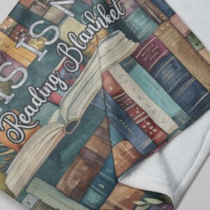 May include: A cozy reading blanket featuring a design of stacked books in various colors. The blanket has the text "THIS IS MY Reading Blanket" in a decorative font. The blanket is folded over, revealing a white, soft underside.