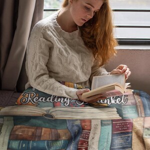 May include: A cozy reading blanket featuring a design of stacked books in various colors. The blanket has the words "Reading Blanket" printed on it. The person in the image is wearing a cream-colored sweater and reading a book.