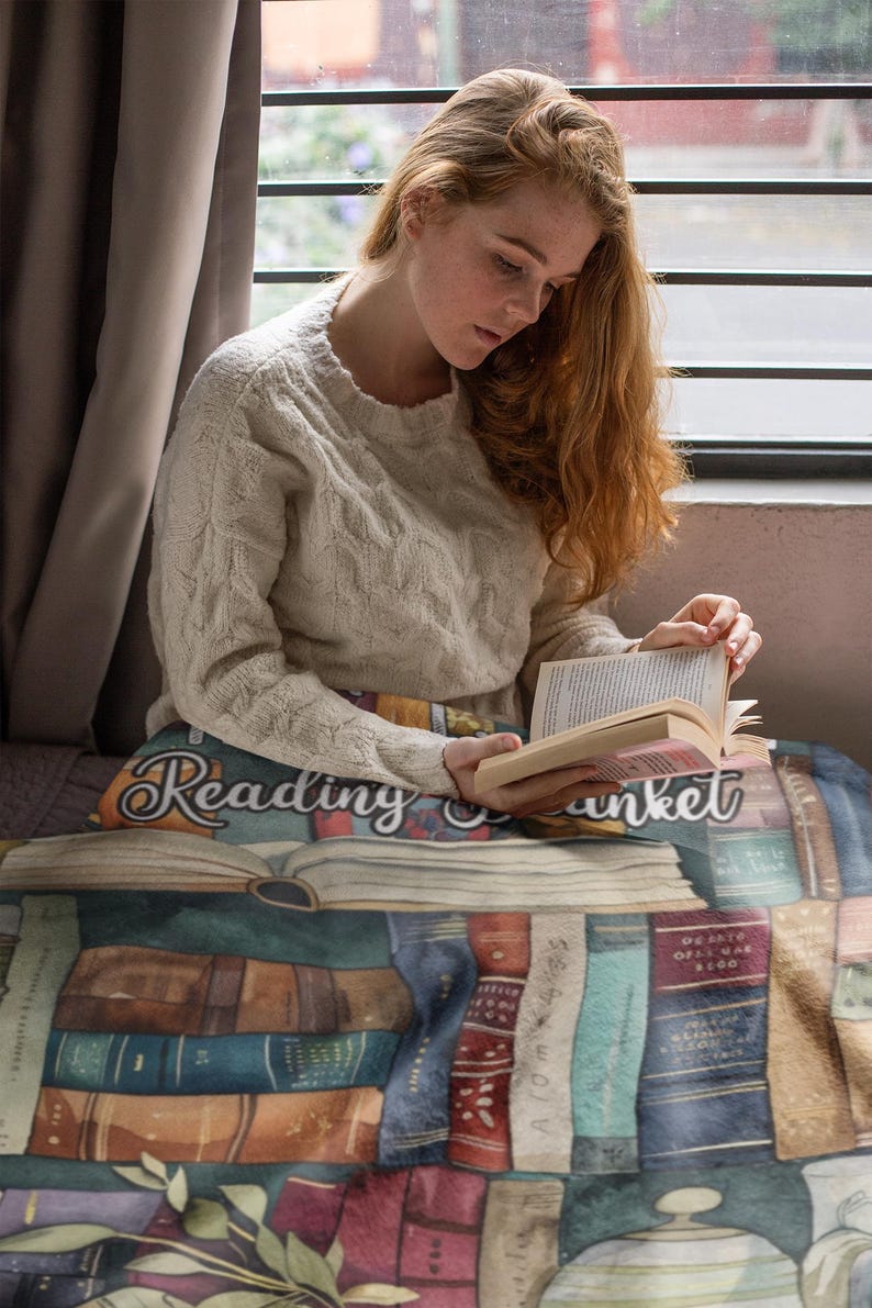 May include: A cozy reading blanket featuring a design of stacked books in various colors. The blanket has the words "Reading Blanket" printed on it. The person in the image is wearing a cream-colored sweater and reading a book.