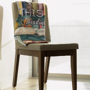 May include: A decorative throw blanket draped over a chair. The blanket features a watercolor design of books, a coffee mug, and the words "THIS READING". The chair has a gray upholstered seat and back with dark brown wooden legs.