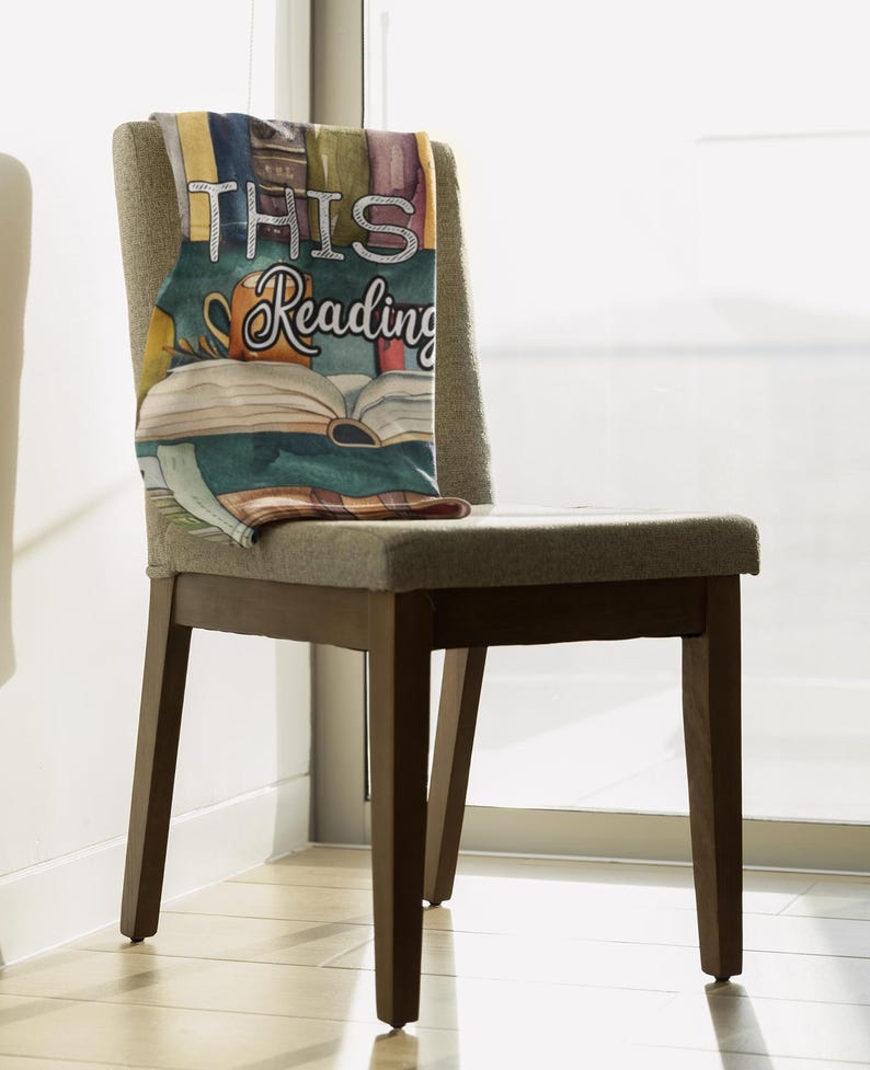 May include: A decorative throw blanket draped over a chair. The blanket features a watercolor design of books, a coffee mug, and the words "THIS READING". The chair has a gray upholstered seat and back with dark brown wooden legs.
