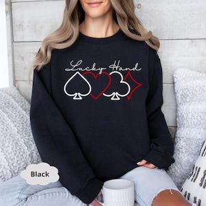 May include: A black sweatshirt with the text "Lucky Hand" and a graphic of four playing card suits: a spade, a heart, a club, and a diamond.