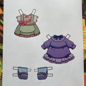 May include: Paper doll clothing set featuring a green and pink dress, a purple dress, and two blue purses. The green dress has a pink bow and heart detail. The purple dress has a blue bow and ruffle details. The text "Paper Dolls By Mila" is at the bottom.