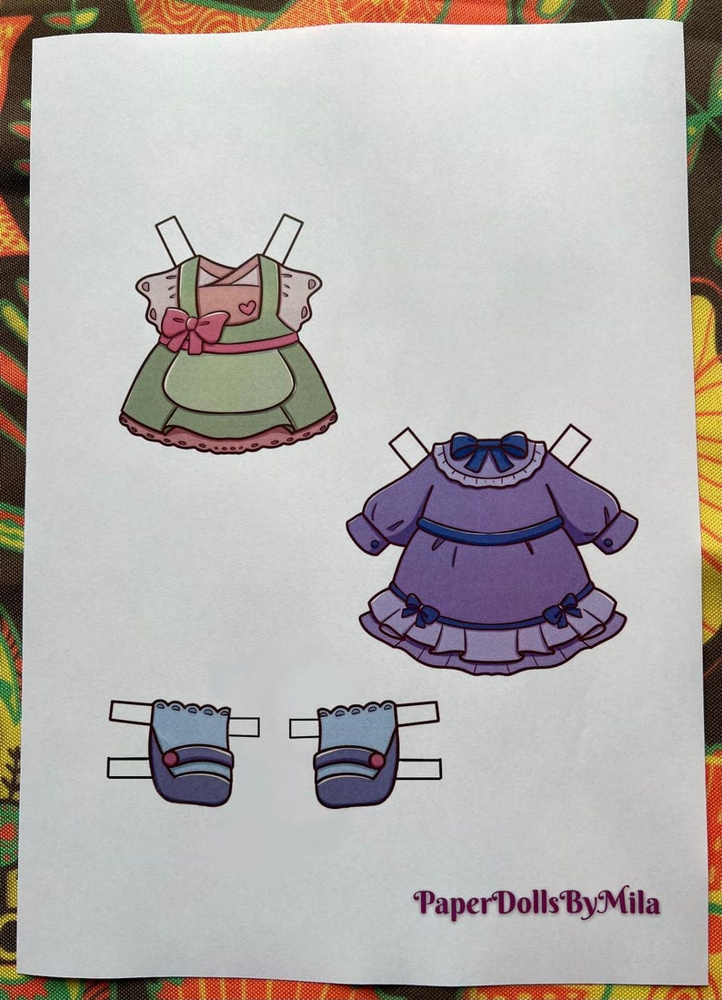 May include: Paper doll clothing set featuring a green and pink dress, a purple dress, and two blue purses. The green dress has a pink bow and heart detail. The purple dress has a blue bow and ruffle details. The text "Paper Dolls By Mila" is at the bottom.