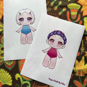 May include: Two paper dolls are depicted, each with a unique hairstyle and swimsuit. One doll has white hair and a blue swimsuit, while the other has purple hair and a pink swimsuit. The text "Paper Dolls By Mila" is visible on the bottom right.