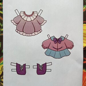 May include: A sheet of paper with three paper doll outfits and two pairs of shoes. The outfits include a pink dress with a white collar, a pink jacket with a blue skirt, and two pairs of purple shoes. The text "Paper Dolls By Mila" is in the bottom right corner.