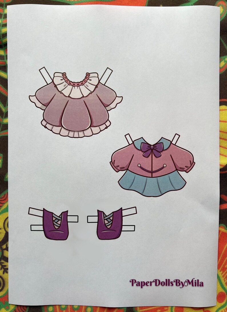 May include: A sheet of paper with three paper doll outfits and two pairs of shoes. The outfits include a pink dress with a white collar, a pink jacket with a blue skirt, and two pairs of purple shoes. The text "Paper Dolls By Mila" is in the bottom right corner.