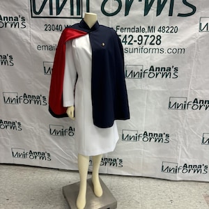 May include: A white medical uniform with a navy blue and red cape. The uniform is on a mannequin and the background is a white wall with the text "Anna's Uniforms" in green letters.