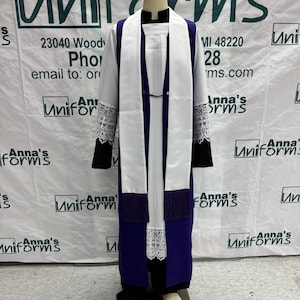 May include: A purple and white liturgical robe with lace trim and a white stole. The robe is on a mannequin and the background is a white sign with the text "Anna's Uniforms" and contact information.
