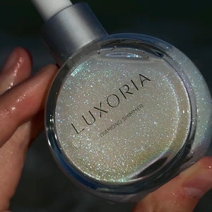 Puede incluir: Una botella de vidrio transparente con una tapa plateada y un gotero blanco. La botella contiene un líquido brillante e iridiscente con el texto "LUXORIA DIAMOND SHIMMER" en la etiqueta.