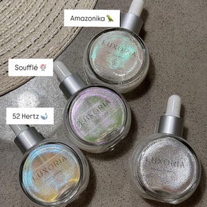 May include: Four clear glass bottles with silver droppers and iridescent glitter inside. The bottles are labeled "Luxoria" and have different colored glitter: green, purple, gold, and silver. The bottles are labeled with names: Amazonika, Souffle, 52 Hertz, and Polaris.