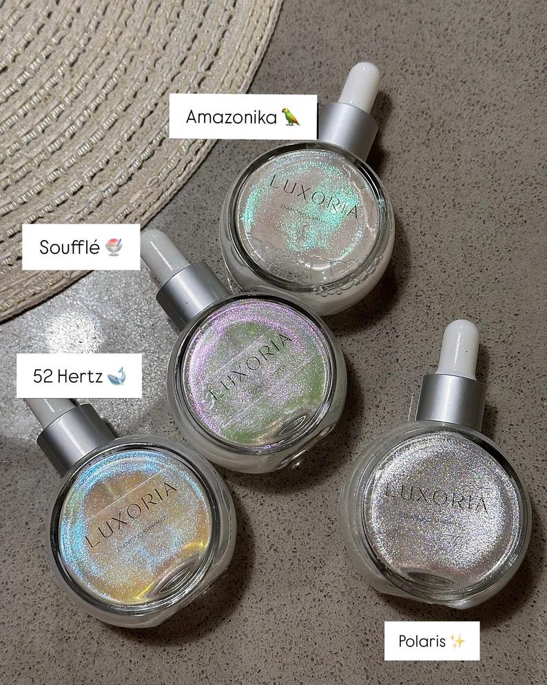 May include: Four clear glass bottles with silver droppers and iridescent glitter inside. The bottles are labeled "Luxoria" and have different colored glitter: green, purple, gold, and silver. The bottles are labeled with names: Amazonika, Souffle, 52 Hertz, and Polaris.