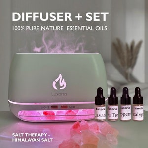 Op de afbeelding: Een groene diffuser met een zoutlampbasis en vier kleine flesjes etherische oliën. De diffuser heeft een licht- en mistinstelling. De flesjes zijn gelabeld met "Luxoria Lavan a", "Luxoria Tea Tre", "Luxoria Pepperm" en "Luxoria Calypt". De tekst "DIFFUSER + SET 100% PURE NATURE ESSENTIAL OILS" staat bovenaan de afbeelding. De tekst "SALT THERAPY - HIMALAYAN SALT" staat onderaan de afbeelding.