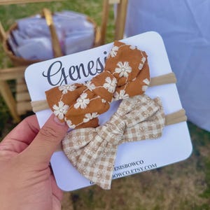 Puede incluir: Dos diademas para bebés con lazos de tela. Un lazo es marrón con bordado floral blanco, y el otro es de cuadros beige. Las diademas están sobre una tarjeta blanca con el nombre "Genesis" y el sitio web "esisbowco.etsy.com".