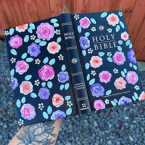 Könnte beinhalten: Geöffnete Heilige Bibel mit einem schwarzen Einband, verziert mit farbenfrohen Blumenmustern. Der Einband zeigt rosa, lila und orangefarbene Rosen, weiße Akzente und den Text "Holy Bible" in goldener Schrift. Der Buchrücken trägt die Aufschrift "Holy Bible" und "English Standard Version".