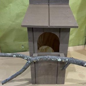 May include: A brown wooden birdhouse with a small opening for birds to enter. A branch is placed in front of the birdhouse.