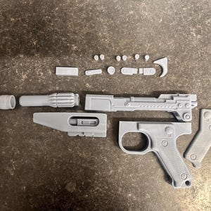 May include: A collection of unpainted grey resin components for a model blaster pistol. The set includes a main barrel, a handle with a trigger guard, a separate grip, and various small mechanical parts, all laid out in a disassembled state.
