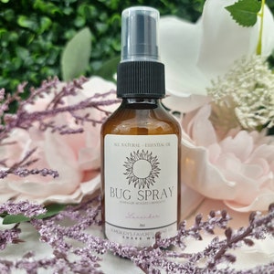 May include: A brown glass bottle with a black spray top filled with bug spray. The label is white with a sun graphic and the text "All Natural - Essential Oil Bug Spray Lavender 200 ml.  Shady Lady's Favourite Scent Co. Shake Well."