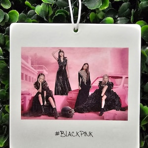 May include: A white ceramic car air freshener with a photo of four women in black dresses standing in front of a pink car. The text '#BLACKPINK' is printed below the photo.