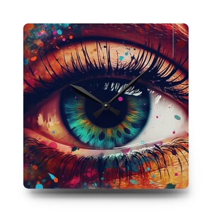 Puede incluir: Un reloj cuadrado con un diseño de ojo abstracto colorido. El ojo es azul, verde y amarillo con pestañas negras y una aguja de reloj negra.