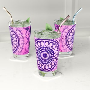 May include: Three clear glass cups with pink and purple mandala designs and straws. The cups are filled with ice and a green leafy garnish.