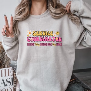 May include: A light beige sweatshirt featuring the text "SERVICE COORDINATOR" in a retro font, with flower and smiley face graphics. The text "HELPING tiny HUMANS MAKE big MOVES" is below in pink. The sweatshirt is a casual style.