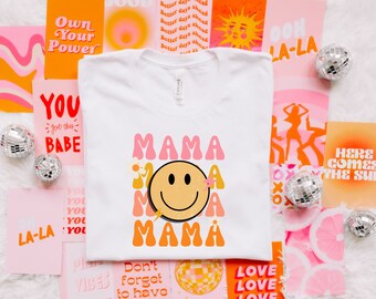 Könnte beinhalten: Weißes T-Shirt mit einem pinkfarbenen, orangen und gelben Grafikdesign, auf dem "Mama" mit einem Smiley in der Mitte steht. Das Design ist von pinkfarbenen und orangen Blumen umgeben.