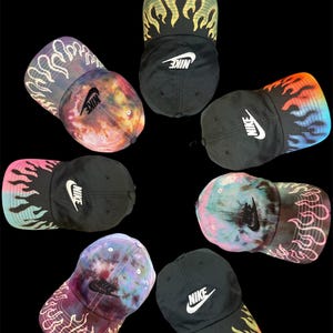 May include: A collection of black baseball caps with the Nike logo in white. The caps feature colorful tie-dye patterns and flame designs in various colors, including yellow, orange, and blue. The caps are arranged in a circular pattern against a black background.