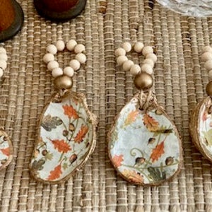 May include: Six fall-themed napkin rings made of oyster shells with a white background and a pattern of orange, brown, and yellow leaves. Each ring is decorated with a strand of wooden beads and a gold accent.