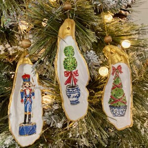 May include: Three oyster shell Christmas ornaments with gold trim. One ornament features a nutcracker, another a topiary tree, and the last a Christmas tree.