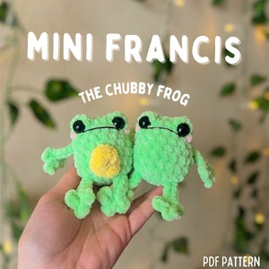 May include: Two green crocheted frog plushies with black eyes and pink cheeks. The frog on the left has a yellow pom-pom on its belly. The plushies are held in a hand. The text "MINI FRANCIS THE CHUBBY FROG PDF PATTERN" is at the top of the image.