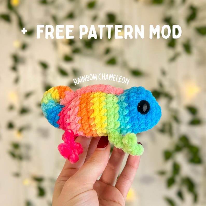 Op de afbeelding: Een handgemaakte gehaakte kameleon in regenboogkleuren. Het pluche speelgoed heeft een opgerolde staart en kleine pootjes. De afbeelding bevat de tekst "+ FREE PATTERN MOD" en "RAINBOW CHAMELEON".