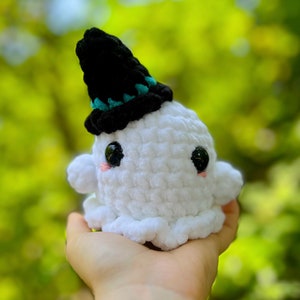 May include: A white crocheted ghost wearing a black witch hat with a teal trim. The ghost has black eyes and pink cheeks.
