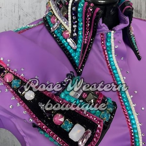 May include: Purple show shirt with black, pink, and turquoise embellishments. The shirt has a pointed collar with rhinestones and a large, decorative design on the left sleeve. The text "Rose Western Boutique" is visible on the shirt.