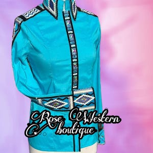 Peut inclure: Une chemise turquoise à manches longues avec des ornements de perles complexes en noir, blanc et bleu. La chemise présente un col haut et des motifs décoratifs sur le devant et les poignets. Le texte "Rose CE Western boutique" est visible.