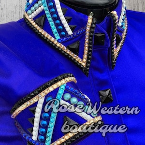May include: A royal blue satin shirt with a black and white geometric design embellished with rhinestones and blue beads. The shirt has a collar and long sleeves. The text "Rose Western Boutique" is visible on the shirt.